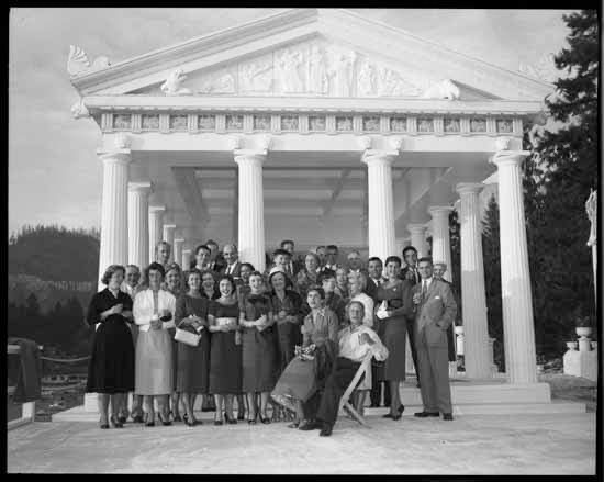 A black and white photo of a group of people standing in front of a replica of the Parthenon, with trees in the background. 